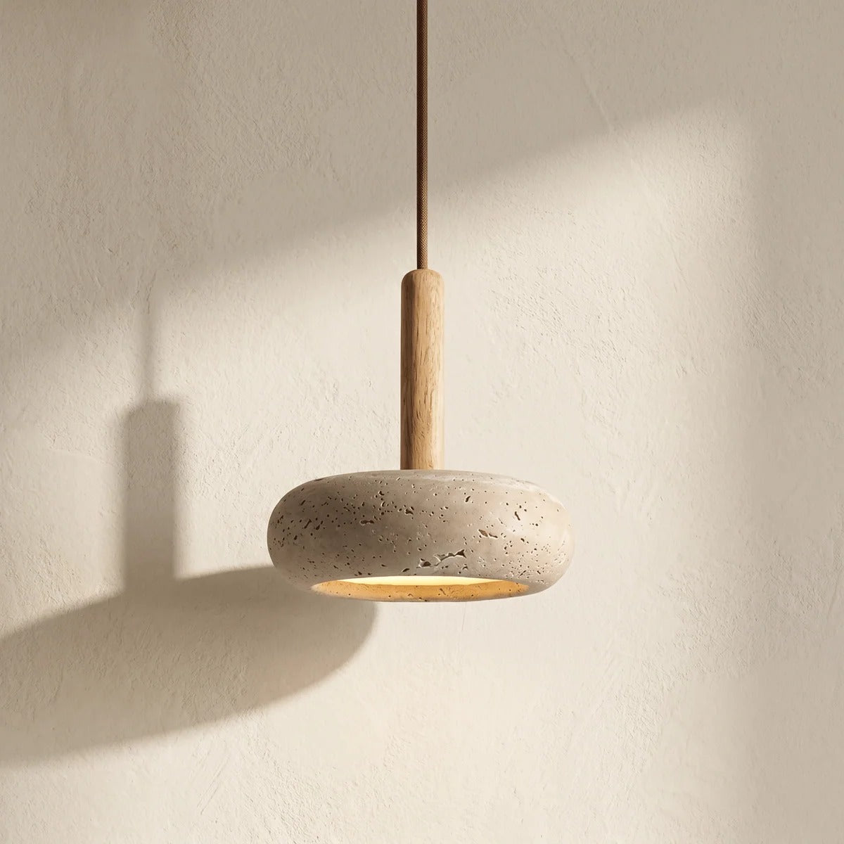 Mila travertine pebble pendant light with a light wood stem, glowing against a cream wall