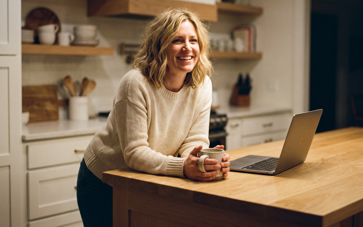 Woman smiling with a coffee mug and laptop at a light oak kitchen island in a warm modern farmhouse kitchen in the evening